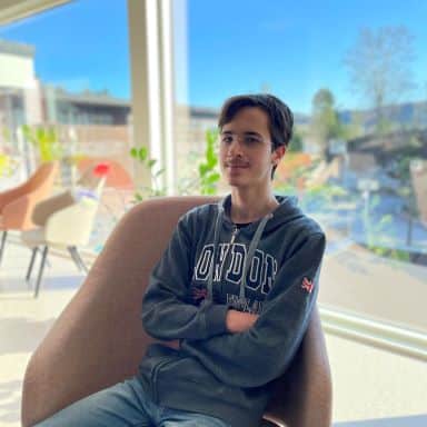 En person med kort, brunt hår, mørk "London"-hettegenser og jeans sitter i en rosa stol ved et stort vindu med planter og utemiljø.