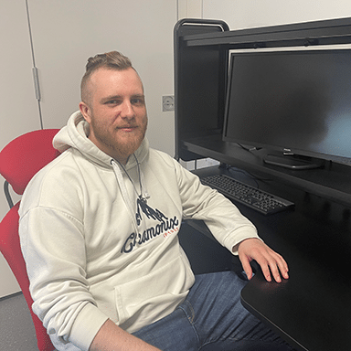 En mann med lyst skjegg og lys hettegenser sitter ved et svart skrivebord foran en dataskjerm og et tastatur i et kontormiljø.