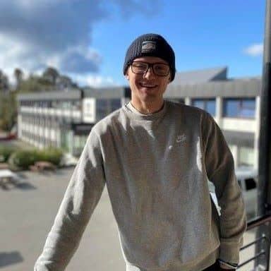 En person med briller, grå genser og mørk lue står på en utendørs balkong med bygninger og en delvis overskyet himmel i bakgrunnen.