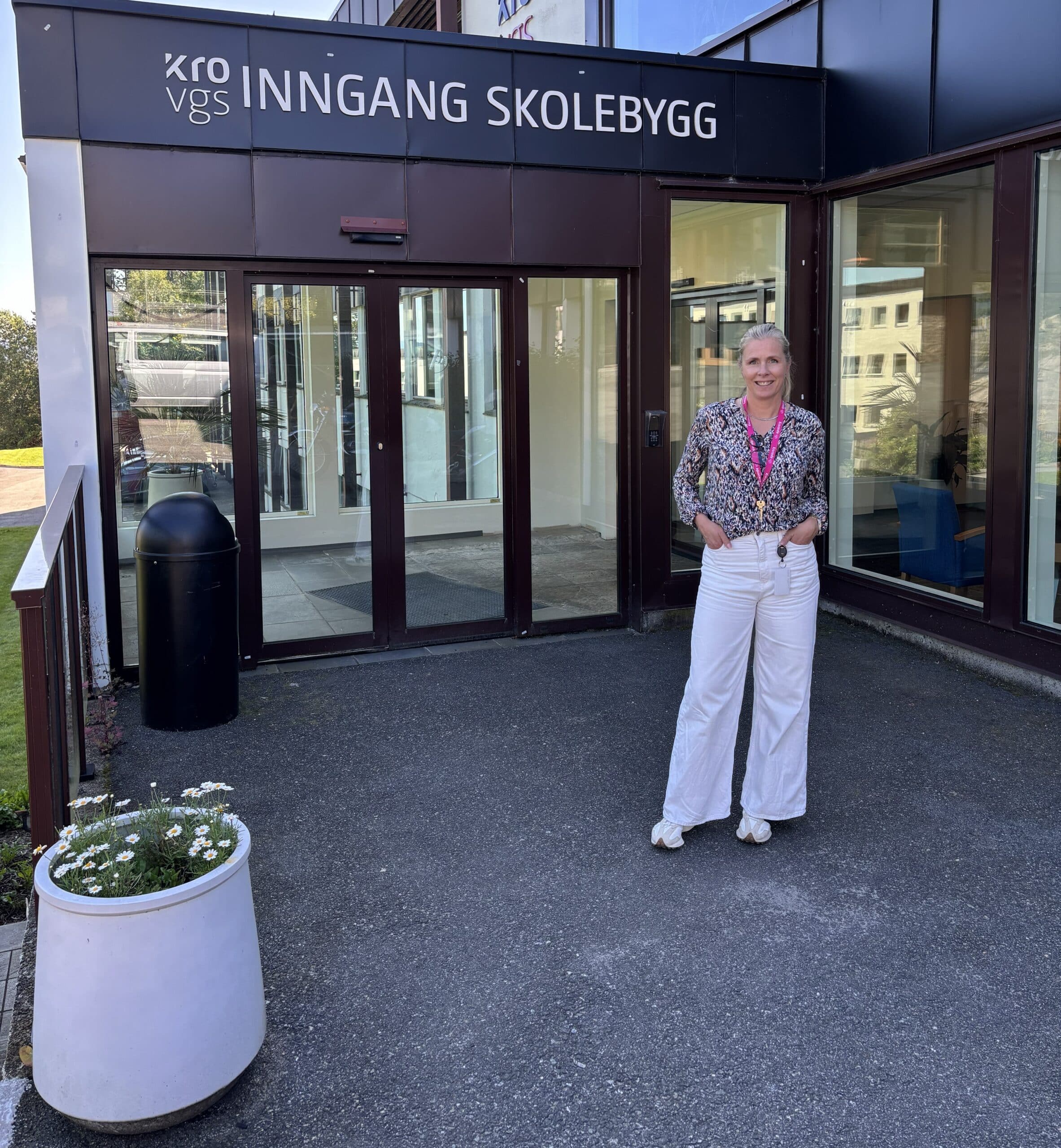En person står utenfor inngangen til et skolebygg med glassdører, under et skilt der det står «INNGANG SKOLEBYGG.».