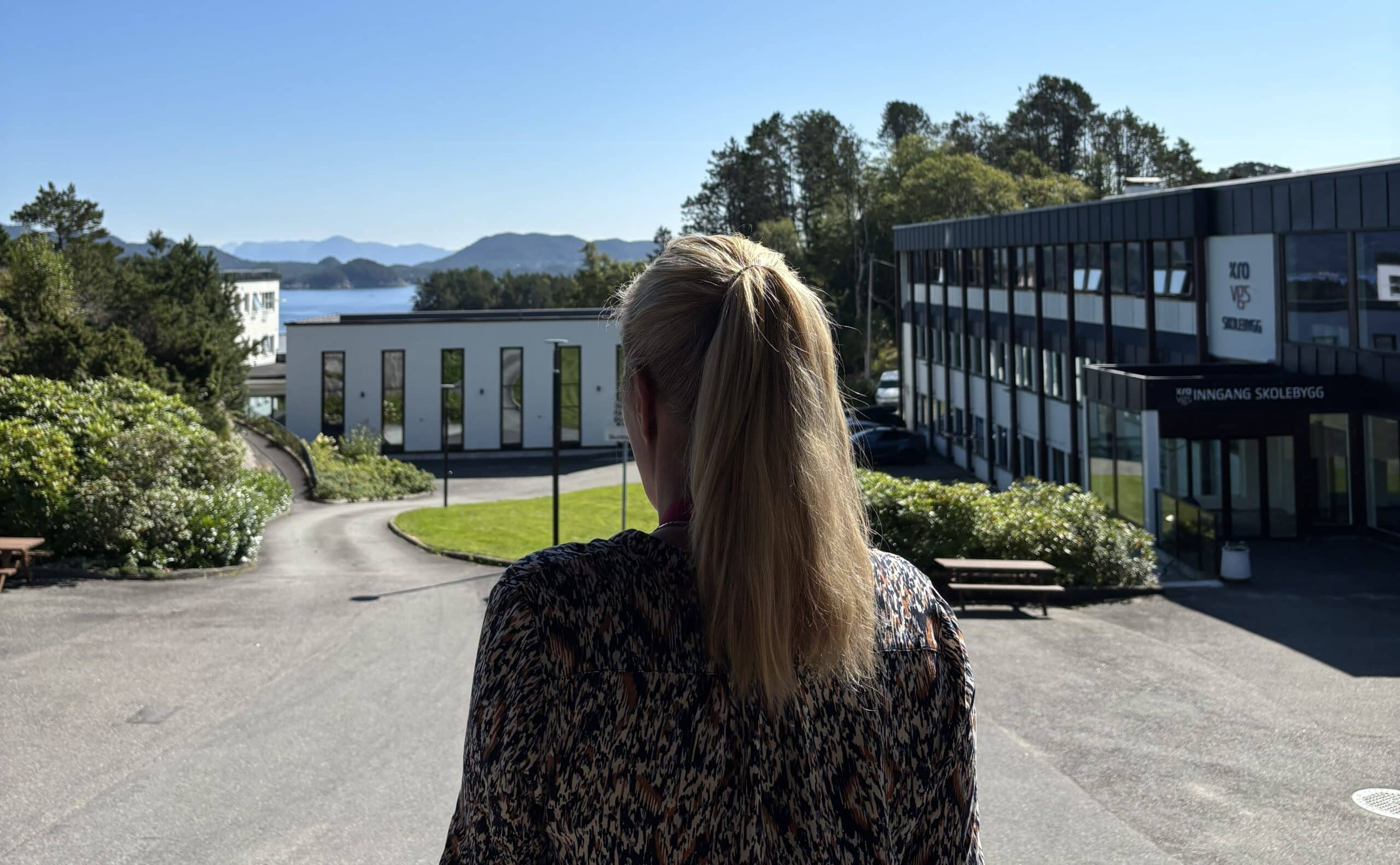 En person med blondt hår i hestehale står ute, med utsikt mot moderne bygninger og et naturskjønt landskap med fjell og vann i bakgrunnen.