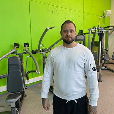 En mann i hvit langermet skjorte står i et treningsstudio med grønne vegger, omgitt av ulike treningsapparater.