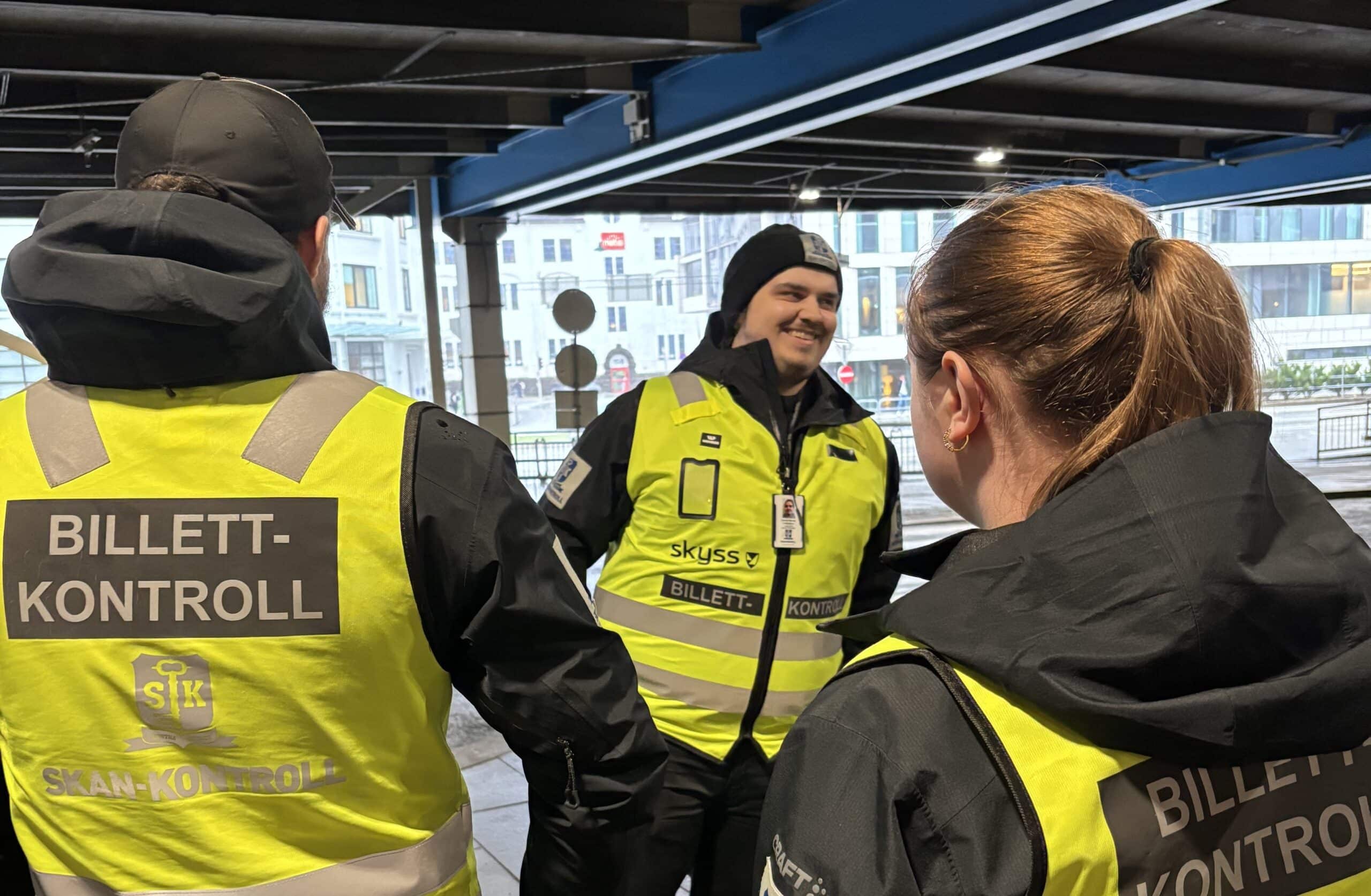 Tre billettkontrollører i gule vester med påskriften "Billettkontroll" står sammen og snakker sammen på en tog- eller trikkestasjon.