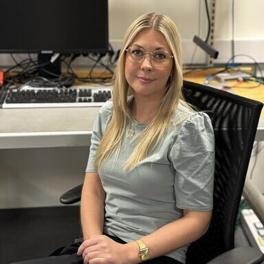 En kvinne med langt blondt hår og briller sitter ved et skrivebord på et kontor, med datautstyr og kabler synlig i bakgrunnen.