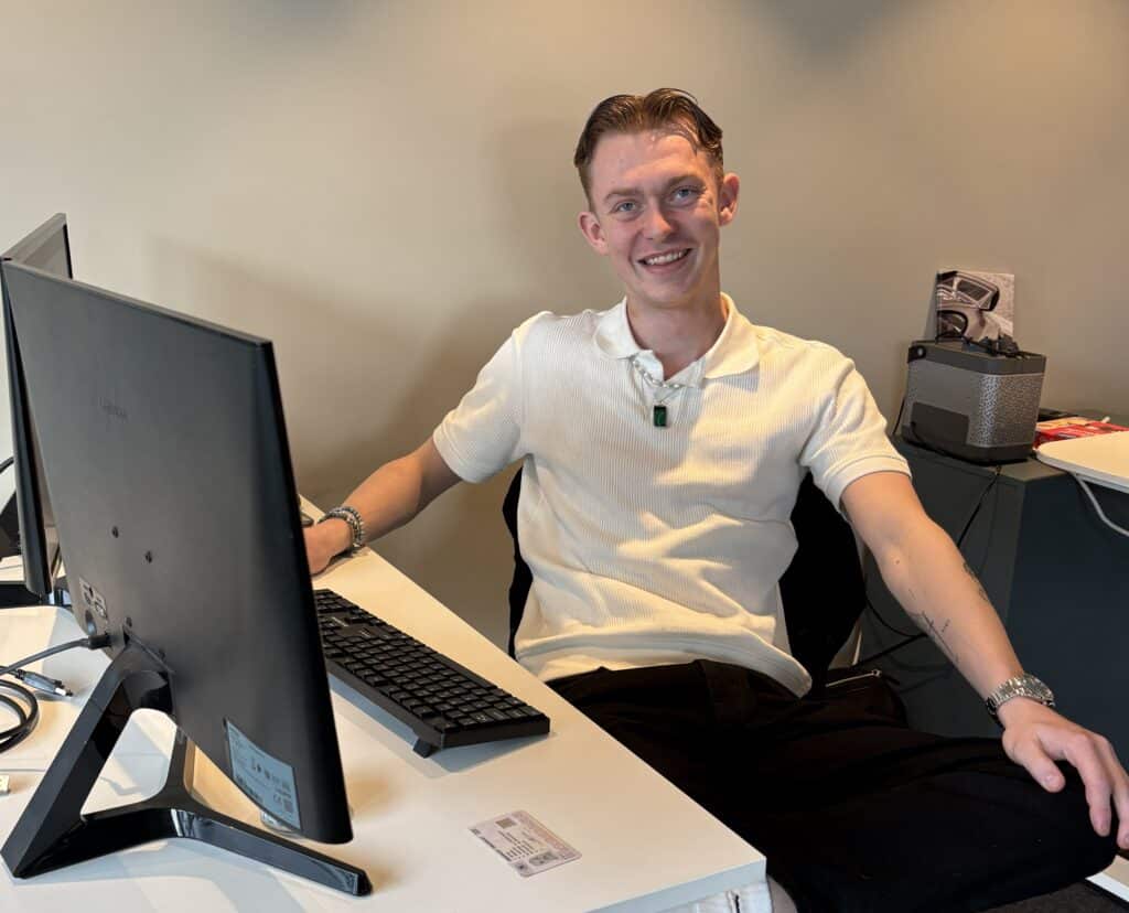 Sebastian praksis2026 (3)_red En smilende person i hvit poloskjorte sitter ved et skrivebord med datamaskin og tastatur i et kontormiljø.