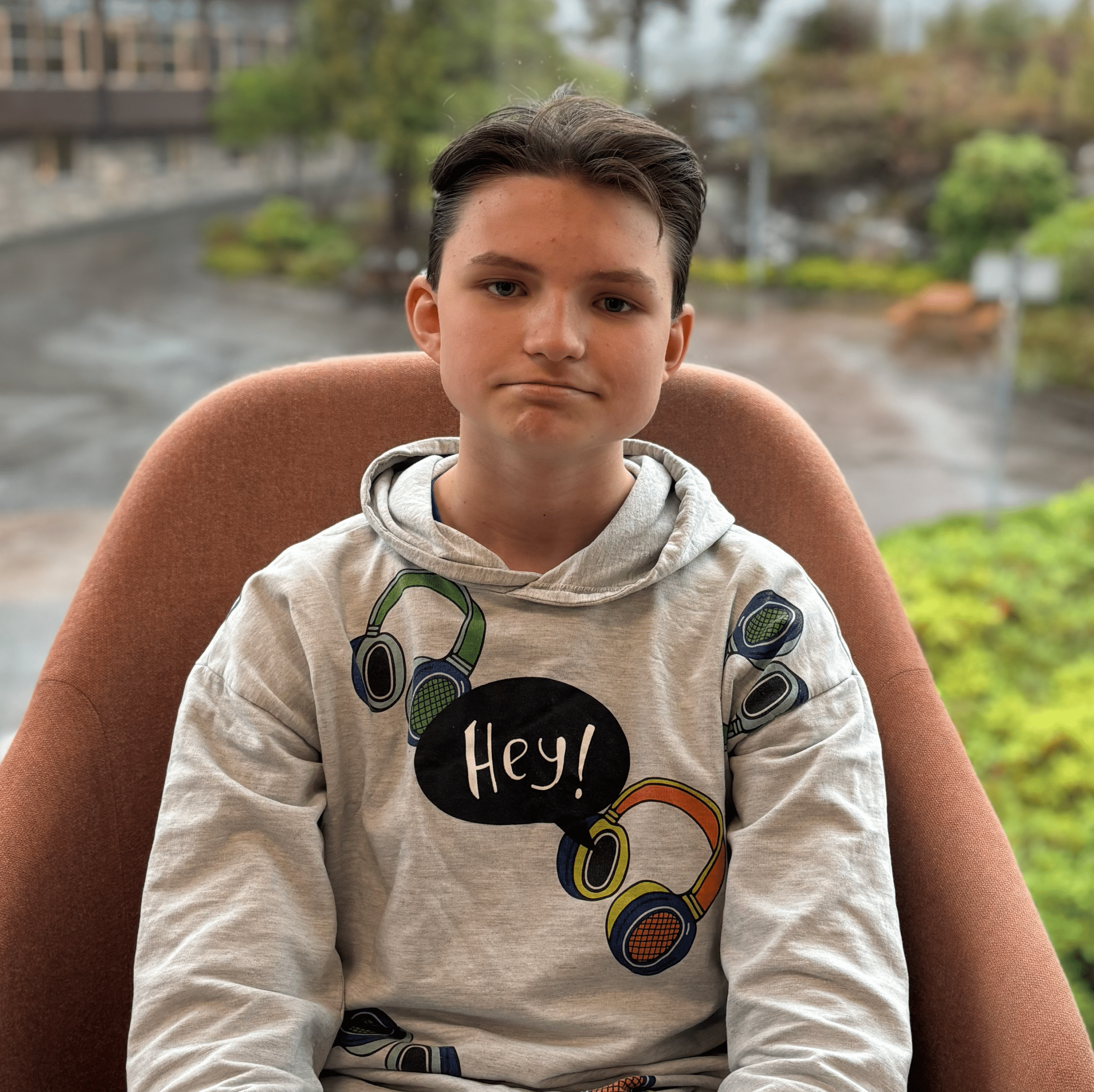 En ung person med kort hår sitter i en stol og har på seg en hettegenser med fargerike hodetelefoner og ordet «Hey!» på. Gjennom vinduet bak vedkommende ser man grønne omgivelser og en bygning.