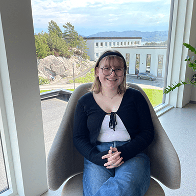 Synneva-Alexandria_2024_web En person med briller og pannebånd sitter i en moderne stol ved et stort vindu med utsikt til trær, steiner og bygninger utenfor.