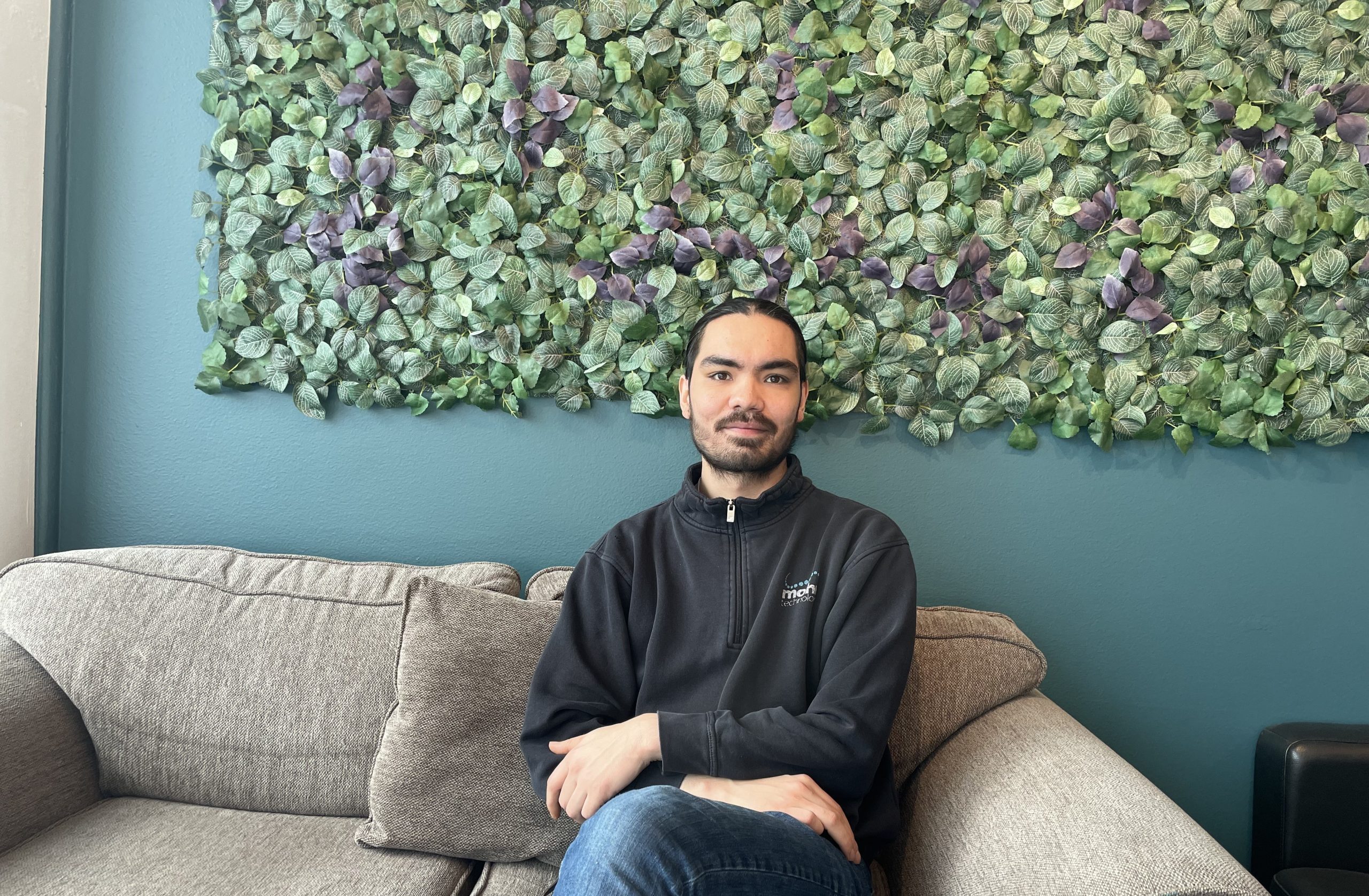 En person med mørkt hår og skjegg sitter i en grå sofa foran en bladgrønn veggdekorasjon, iført svart jakke og blå jeans.