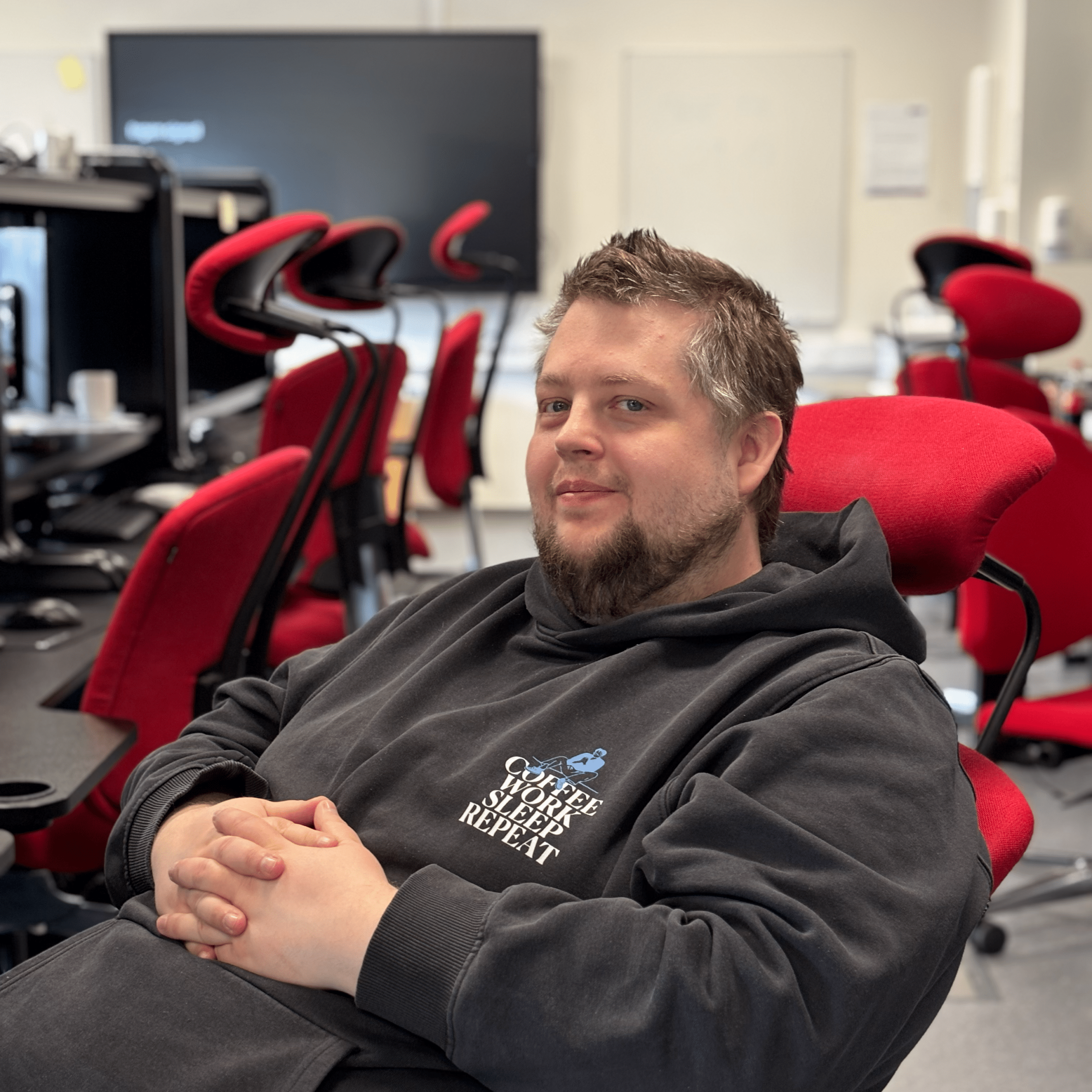 En mann med kort hår og skjegg sitter i en rød kontorstol, kledd i en mørk hettegenser, i et rom med røde stoler, skrivebord og dataskjermer.