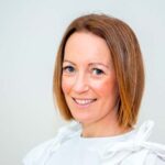 En kvinne med rett, skulderlangt, lysebrunt hår og blå øyne smiler til kameraet, iført en hvit topp mot en ensfarget, lys bakgrunn.