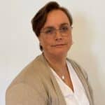 En person med kort, brunt hår, briller, beige kofta og hvit skjorte sitter foran en vanlig, lys bakgrunn og ser inn i kameraet.