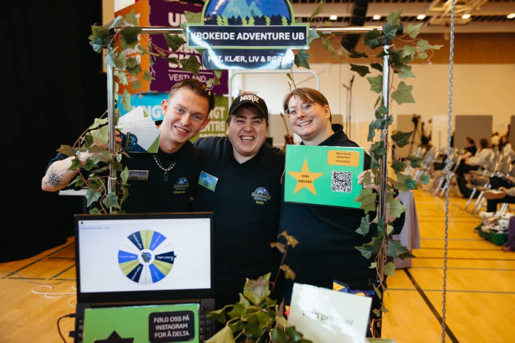 UE 2026 Tre personer står smilende bak en dekorert stand på et arrangement, med en bærbar datamaskin, skilt og pyntegrønt.