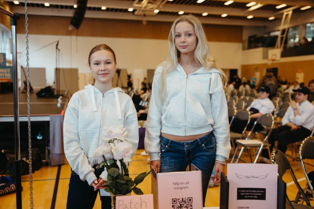 UE 2026 To jenter i lyse hettegensere står innendørs bak et bord med skilt og blomster på et arrangement, med folk sittende i bakgrunnen.