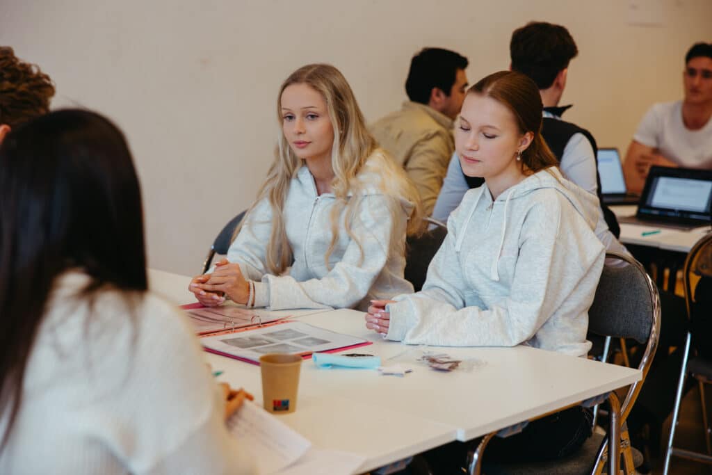 UE 2026 To unge kvinner sitter ved et bord med papirer og en kaffekopp og deltar i en gruppediskusjon i et klasserom, med andre studenter i bakgrunnen.