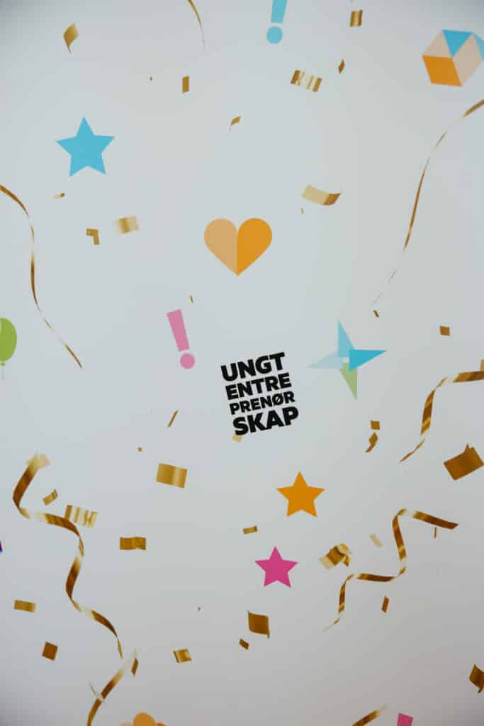 UE 2026 Foto: Fotograf Inger Paulsen Fargerike papirformer, gullbånd og konfetti omkranser den svarte teksten "UNGT ENTREPRENØR SKAP" på hvit bakgrunn.