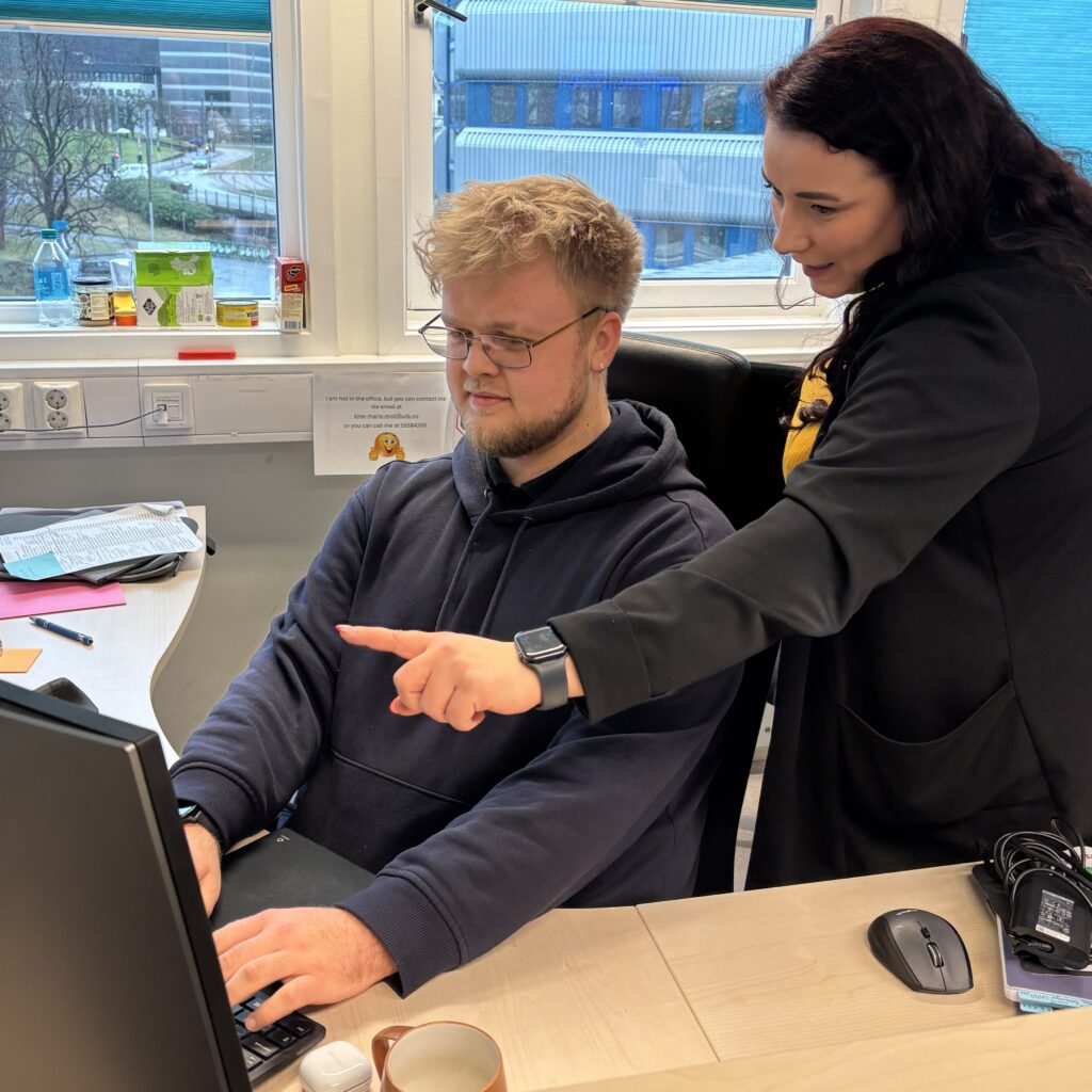 UiB_Kristian 2026 (6)_red En mann sitter ved et skrivebord og skriver på et tastatur, mens en kvinne står ved siden av ham og peker på dataskjermen. Begge er fokusert på skjermen.