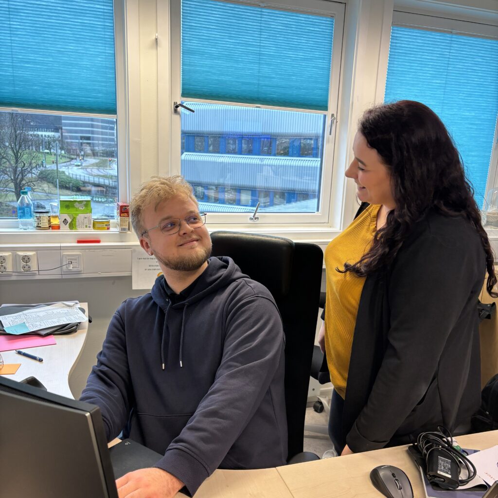 UiB_Kristian 2026 (8)_red En mann sitter ved et skrivebord med en datamaskin og ser opp på en kvinne som står ved siden av ham. Det ser ut til at de fører en samtale i et kontormiljø.