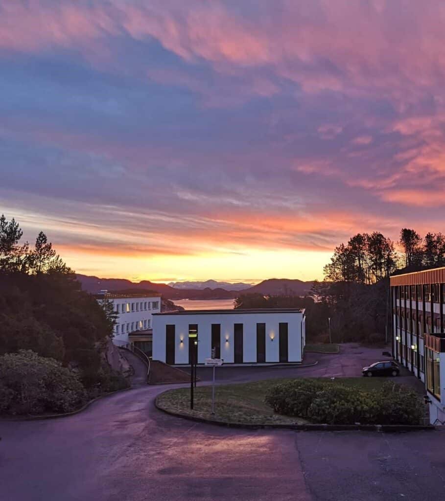 Ukens-bilde-uke-1_2022 En fargerik solnedgangshimmel over moderne bygninger, trær og en asfaltert vei, med fjerne åser og vann i bakgrunnen.