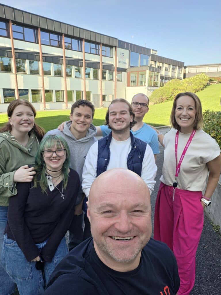 En gruppe på syv personer som poserer for et selfie foran en skolebygning.
