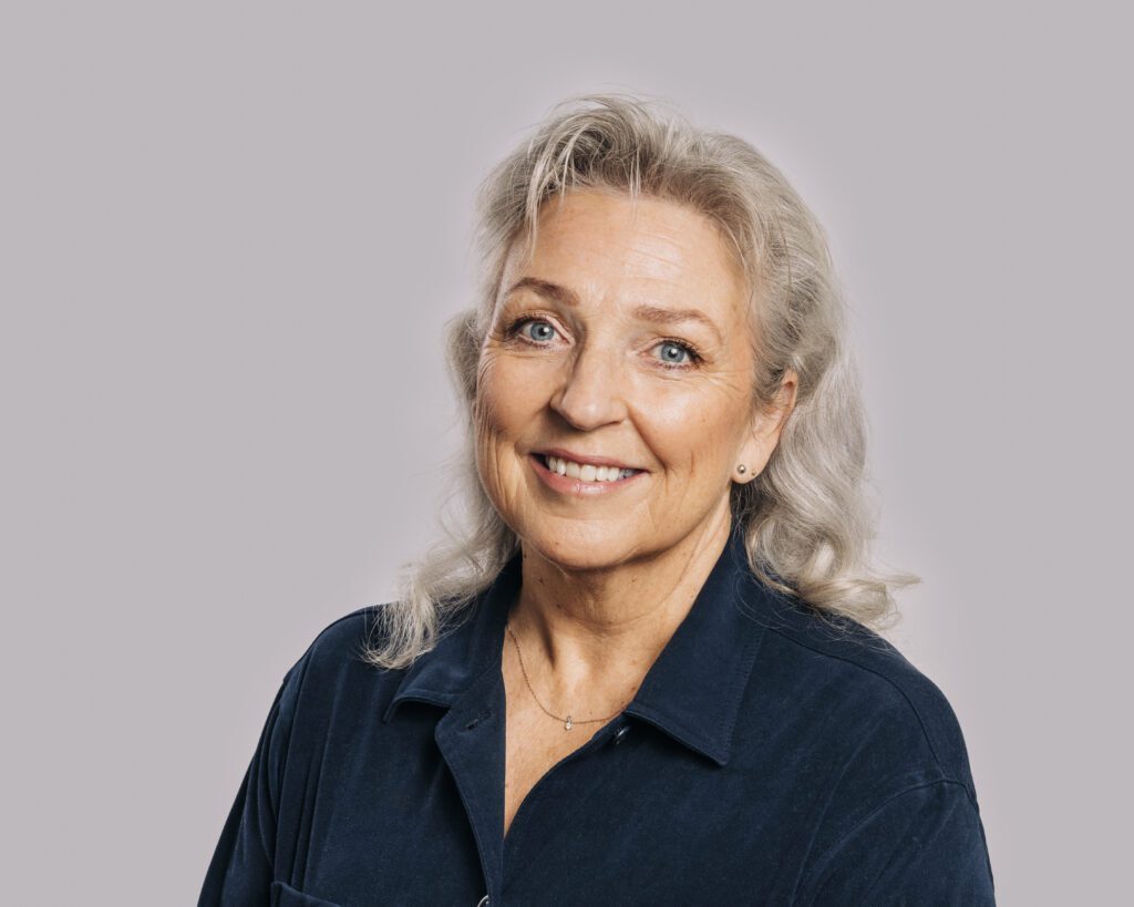 Videre_skolene-5-1024x819 En eldre kvinne med grått hår og blå øyne, iført mørkeblå skjorte, smiler til kameraet mot en vanlig lysegrå bakgrunn.