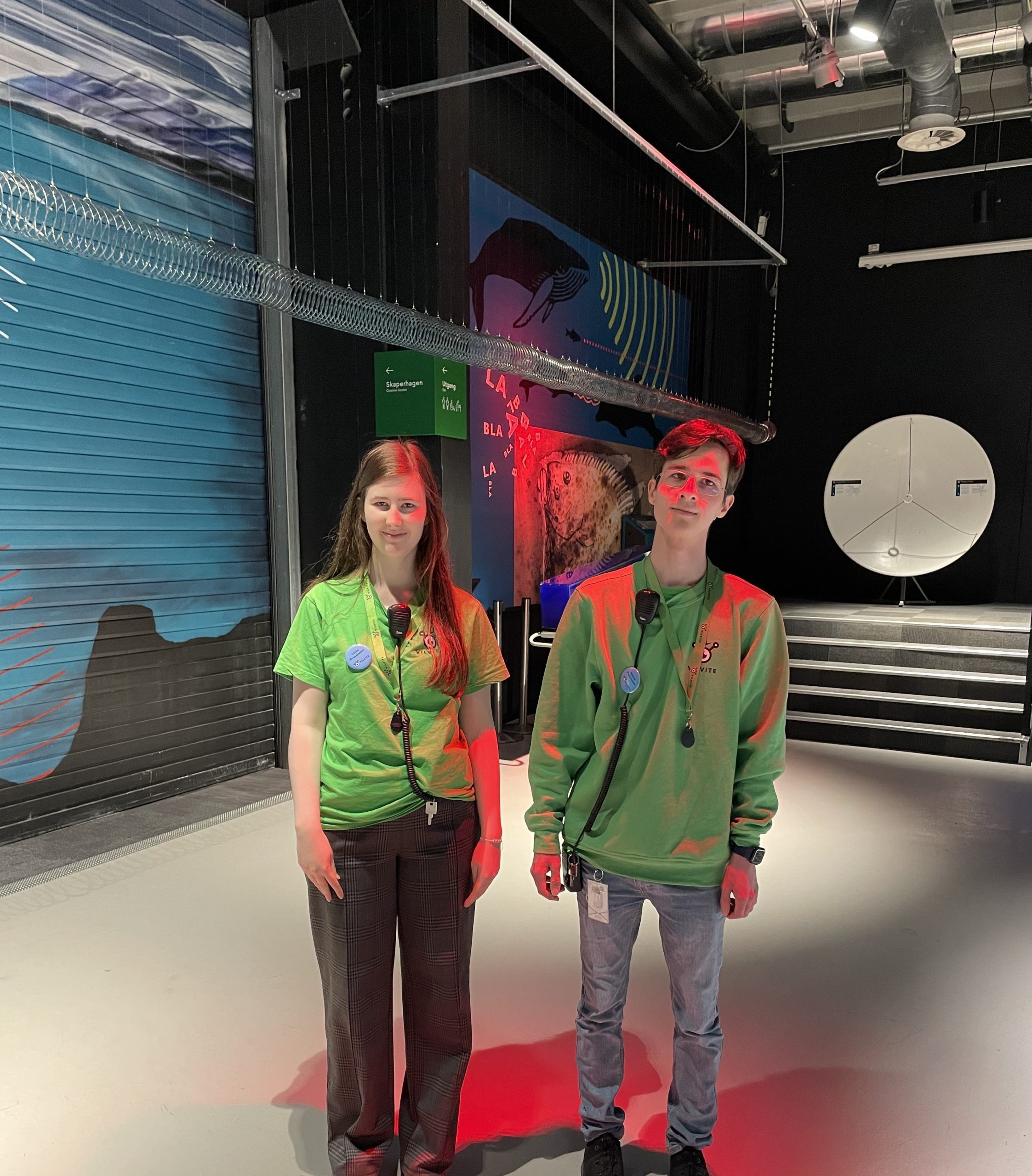 To personer i grønne skjorter og nøkkelbånd står innendørs under sterk belysning, med industrielle takrør og et fargerikt veggmaleri bak seg.