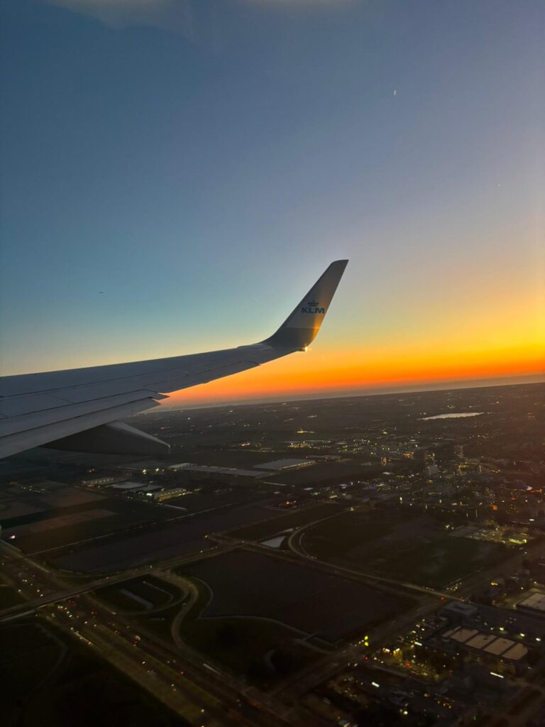 Utsikt fra et flyvindu ved solnedgang, med flyvingen, bylysene nedenfor og en himmel som går fra blått til oransje.