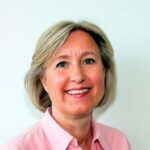 En kvinne med kort, blondt hår, iført en lys rosa skjorte med krage, smiler foran en vanlig hvit bakgrunn.