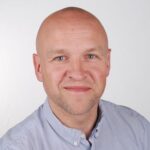 En skallet mann med lys hud, iført lyseblå skjorte, smiler til kameraet mot en hvit bakgrunn.