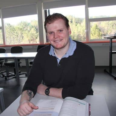 En mann med rødlig hår og skjegg sitter ved en pult i et klasserom og smiler mot kameraet med en oppslått bok foran seg. Vinduer viser utsikten til trær utenfor.