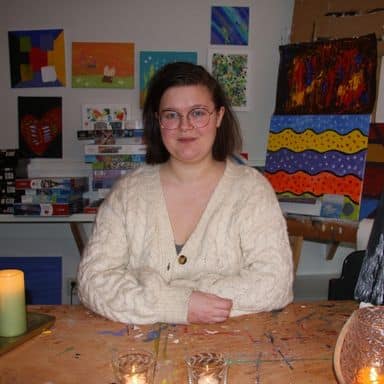 En person med briller og kremfarget genser sitter ved et bord i et kunstatelier, omgitt av fargerike malerier og tente stearinlys.