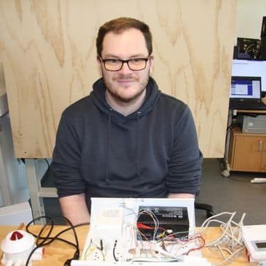 En person med briller og mørk hettegenser sitter ved et skrivebord med diverse elektroniske komponenter og ledninger foran seg.