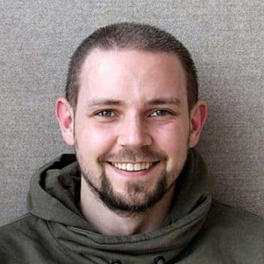 En mann med kort, brunt hår, skjegg og bart smiler til kameraet mens han er iført en grønn hettegenser mot en ensfarget bakgrunn.