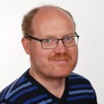 En mann med briller, kort, rødlig hår og rødlig skjegg har på seg en blå- og svartstripet skjorte og ser inn i kameraet mot en vanlig, hvit bakgrunn.