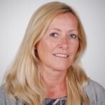 En kvinne med langt, blondt hår, iført en grå cardigan over en mønstret topp, smiler lett foran en ensfarget, lys bakgrunn.