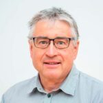 Middelaldrende mann med kort, grått hår, briller og lysegrå skjorte med krage, vendt mot kameraet og smilende mot en vanlig, lys bakgrunn.