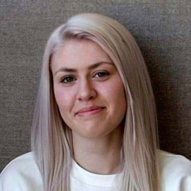 En kvinne med langt, rett, platinablondt hår og et nøytralt uttrykk sitter foran en grå bakgrunn.