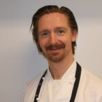 En mann med rødbrunt hår og skjegg, iført hvit kokkefrakk og svart forkle, står mot en ensfarget, lys bakgrunn.