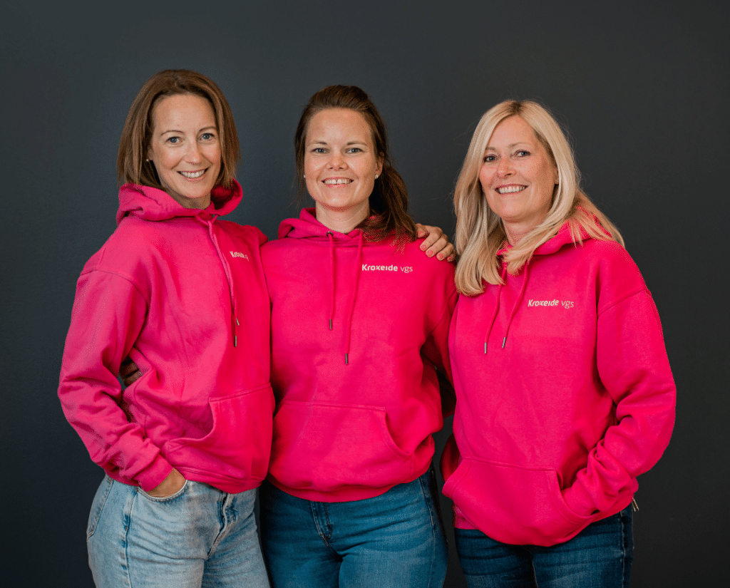 Tre kvinner står side om side, smilende og iført matchende lyserosa hettegensere og jeans mot en ensfarget, mørk bakgrunn.