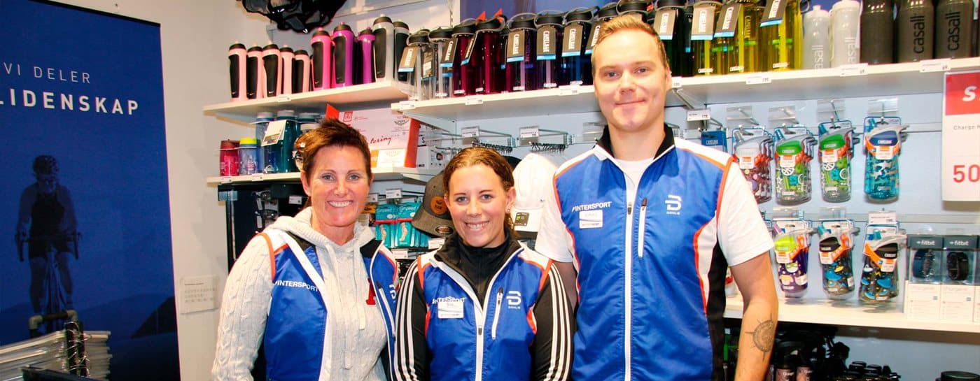 Tre butikkansatte i blå vester står foran hyller med vannflasker og sportsutstyr og smiler til kameraet.