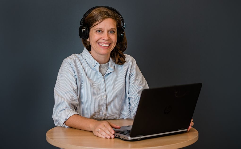 En kvinne med hodetelefoner sitter ved et rundt bord og smiler mot kameraet, med en bærbar datamaskin åpen foran seg mot en enkel, mørk bakgrunn.
