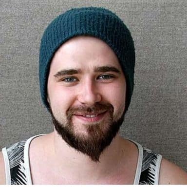 En ung mann med mørkt skjegg og bart, iført blå strikkelue og mønstret tanktop, smiler foran en grå bakgrunn.