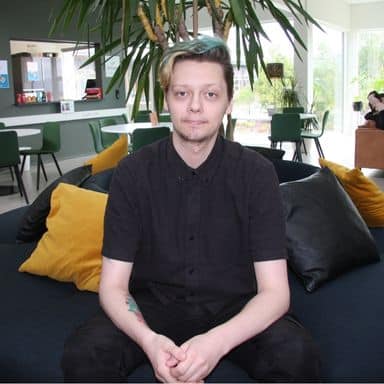 En person med kort, blondt hår og svart skjorte sitter i en mørk sofa med gule og svarte puter; moderne, lyst rom med grønne planter og stoler i bakgrunnen.