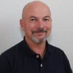 En middelaldrende mann med skallet hode, fippskjegg og bart har på seg en mørk skjorte med krage og smiler til kameraet mot en vanlig lys bakgrunn.