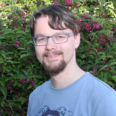 En mann med briller og skjegg står utendørs foran grønt løvverk og rosa blomster, iført en lyseblå T-skjorte og smiler til kameraet.