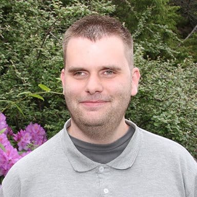 En mann med kort, brunt hår og kortklippet skjegg står ute foran grønne busker og rosa blomster, iført en grå skjorte med krage.