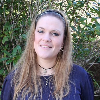 En kvinne med langt, lysebrunt hår, iført marineblå topp og flere lag med halskjeder, står utendørs foran grønne busker.