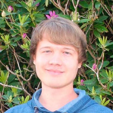 En ung mann med lysebrunt hår og blå hettegenser står foran grønne busker med rosa blomsterknopper.