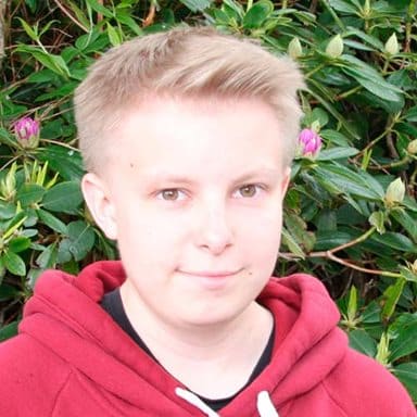 En person med kort, blondt hår og rød hettegenser står foran et grønt bladverk med rosa blomsterknopper.