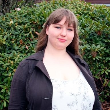 En person med langt brunt hår og mørk jakke over en hvitmønstret overdel står utendørs foran grønne busker.