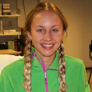 En kvinne med langt, blondt hår i to fletter, iført grønn jakke med glidelås, smiler mens hun sitter innendørs med hyller og bøker i bakgrunnen.